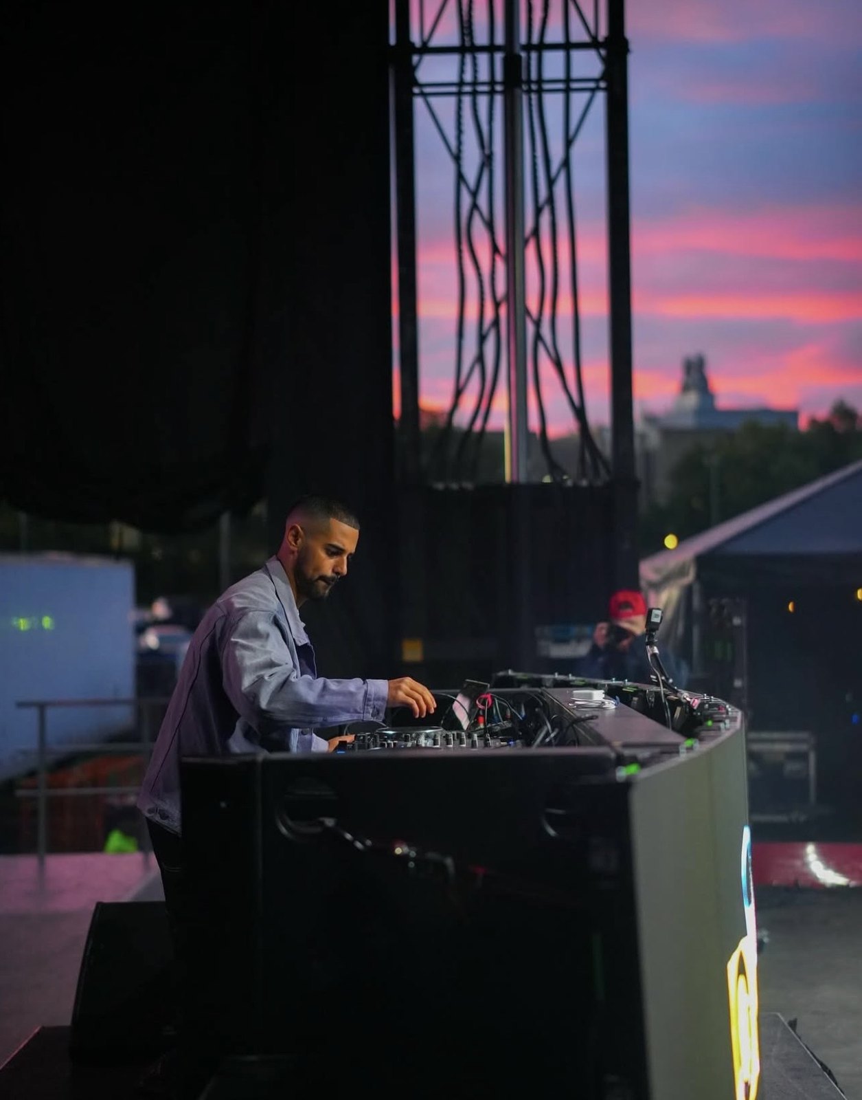 DJ performing on an outdoor stage against a colourful sunset